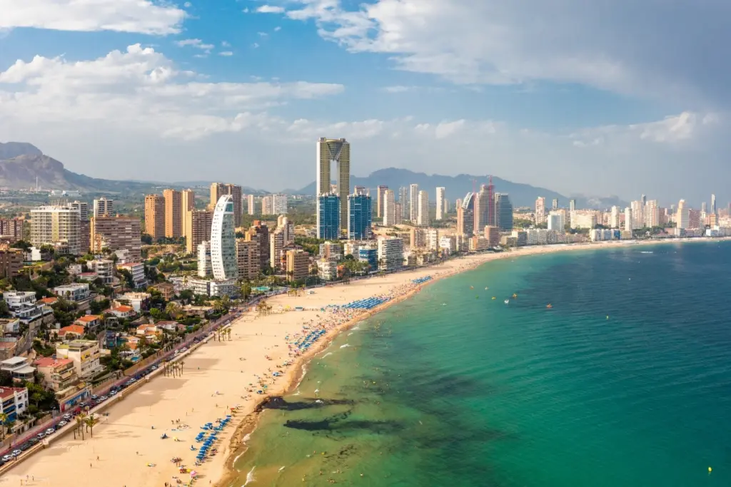 Aerial view of Benidorm's high-rise skyline along a busy sandy beach, with mountains behind the town