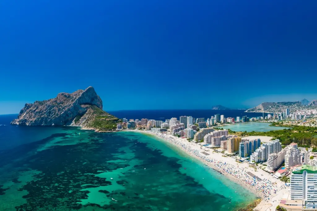 Aerial view of La Fossa beach in Calpe curving beneath the Peñón de Ifach, with Las Salinas salt lake and the town behind