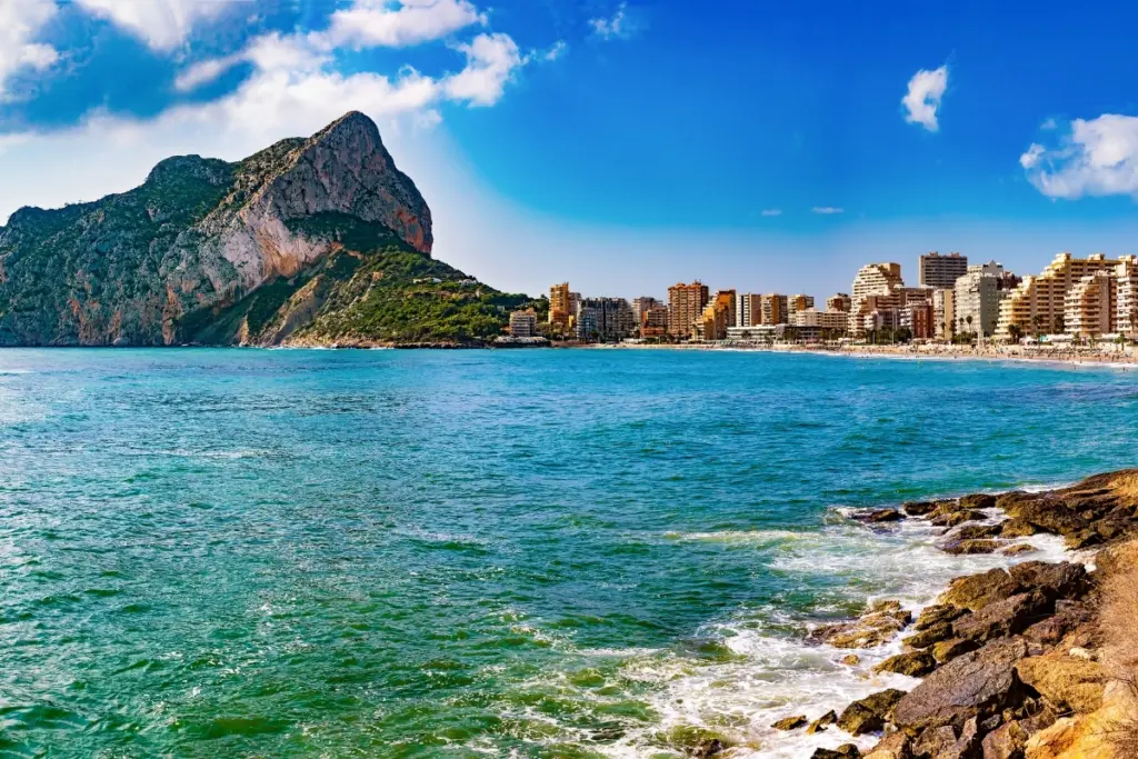 View across Calpe's bay towards the Peñón de Ifach, with the beachfront town curving beneath the rock and a rocky promenade in the foreground